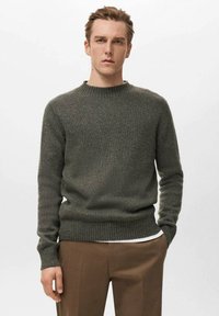 Pull tricoté vert olive avec un col rond et des poignets et une base côtelés. Porté sur une chemise blanche, présentant une apparence douce et texturée.