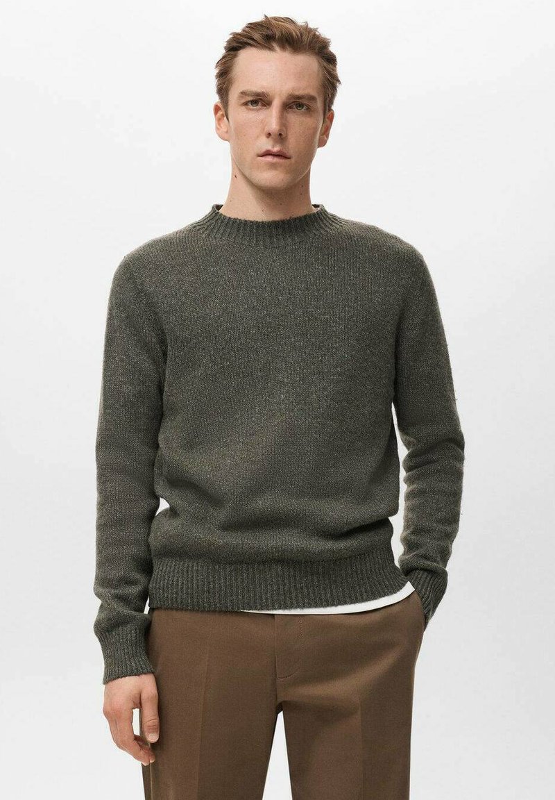 Pull tricoté vert olive avec un col rond et des poignets et une base côtelés. Porté sur une chemise blanche, présentant une apparence douce et texturée.