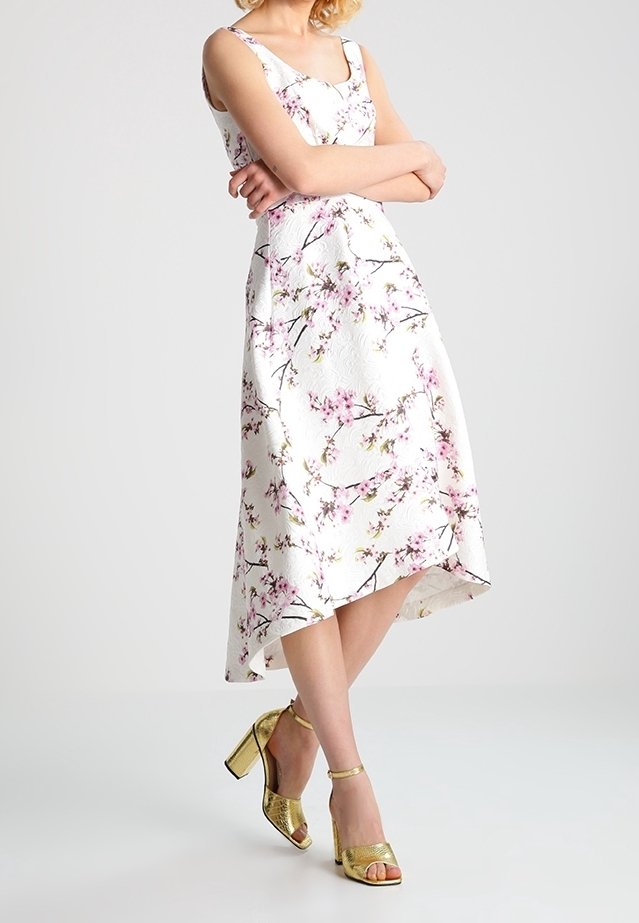 Femme portant une robe midi blanche fleurie sans manches avec des fleurs roses et des sandales à talons hauts dorées, debout les bras croisés.