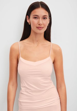 Junge Frau mit langen dunklen Haaren, die ein blass rosa Camisole-Top mit dünnen Trägern vor einem einfarbigen hellen Hintergrund trägt.