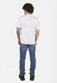 T-shirt bianco a maniche corte, jeans in denim, scarpe sportive nere. La vista posteriore mostra una vestibilità comoda e un design minimalista. Si vedono capelli ricci.