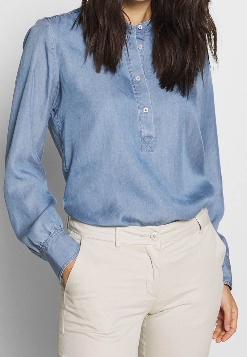 Light blue denim shirt with a button-up collar, two-button placket, and long sleeves. Paired with light-colored, fitted pants.