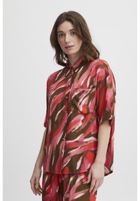Blusa a maniche corte con un motivo astratto multicolore nei toni del rosa e del marrone, dotata di una chiusura con bottoni e di una tasca sul petto.