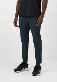 Herre tapered løbebukser i mørk teal, lavet af letvægtsmateriale. Har sidelommer og en mat finish. Sammen med sorte sneakers.