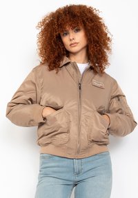 Beige Bomberjacke mit Reißverschluss, gerippten Bündchen und Taschen. Glattes, glänzendes Material mit einem Logo-Patch auf der Brust. Lässiges Design.