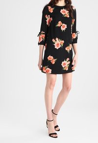 Czarna sukienka w kwiaty z trzema ćwiartkami rękawów, z różowymi i pomarańczowymi kwiatami oraz detalami w postaci wiązań po bokach. Połączona z czarnymi sandałami na obcasie.