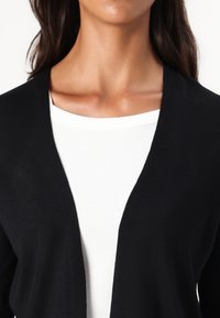 Zwarte cardigan van zacht gebreid materiaal met een diepe V-hals, gelaagd over een witte geribde top, die soepele texturen en strakke lijnen laat zien.