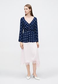 Ναυτικό μπλούζα με λευκές πουά, ρυτιδωμένο μπροστινό μέρος και μακριά μανίκια, συνδυασμένη με φούστα από απαλό ροζ σατέν με δαντελωτό τελείωμα. Λευκά τακούνια.