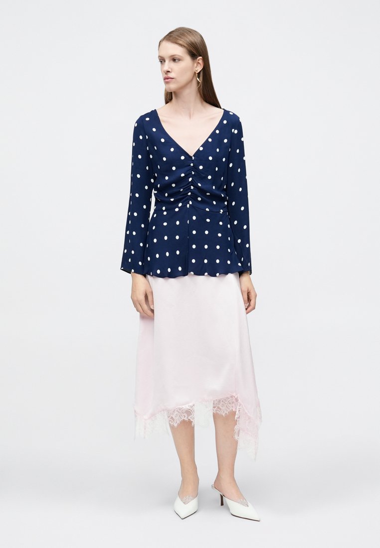 Blouse marine à pois blancs, devant froncé, manches longues, associée à une jupe en satin rose clair avec un ourlet en dentelle. Talons blancs.