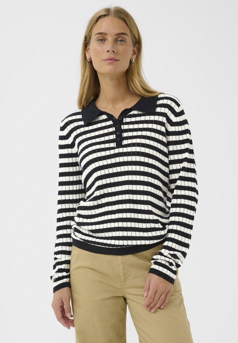 Donna bionda che indossa un maglione polo a righe bianche e nere con maniche lunghe e pantaloni beige, in piedi su uno sfondo bianco.