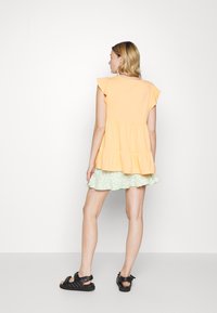 Top peplum em tecido laranja suave com mangas em riste; combinado com uma saia floral verde-claro; sandálias pretas com tiras.