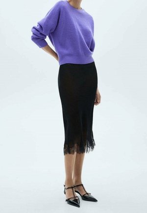 Femme portant un pull violet et une jupe midi noire avec une bordure en dentelle, debout avec une main derrière le dos, portant des talons aiguilles noirs.