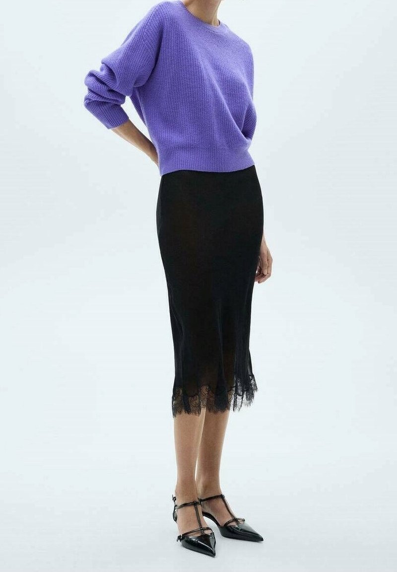 Femme portant un pull violet et une jupe midi noire avec une bordure en dentelle, debout avec une main derrière le dos, portant des talons aiguilles noirs.