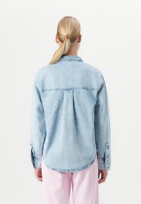Femme blonde avec une queue de cheval portant une veste en jean bleu clair et un pantalon rose, se tenant devant un fond blanc.