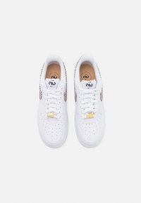 Paire de baskets blanches Nike avec accents imprimé léopard, étiquettes de lacets dorées swoosh et logo noir sur la languette, vues de dessus.