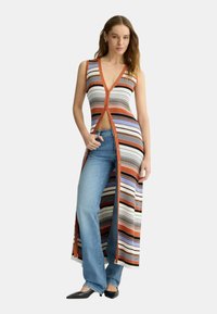 Gestreepte gebreide cardigan jurk in oranje, wit, paars en zwart, met een diepe V-hals, knoopsluiting aan de voorkant en lange zijslits, gecombineerd met flared jeans.