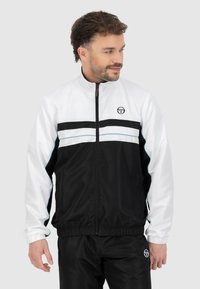 Zwart-wit windbreakerjack met een hoge kraag, elastische manchetten en twee horizontale strepen met een blauwe accent over de borst.