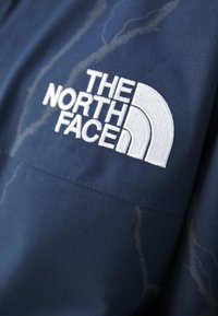 Tmavě modrá látka bundy s jemným světlým mramorováním, s výrazným bílým vyšívaným logem The North Face na hrudi.