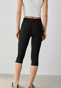 Femme portant un pantalon noir raccourci et ajusté, un haut blanc sans manches et des chaussures beige tissées sans lacets, debout de dos sur un fond uni.