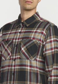 Geruite flanellen shirt in zwart, rood, grijs en geel. Kenmerken zijn een kraag, twee voorzakken en knoopsluiting. De stof heeft een zachte textuur.