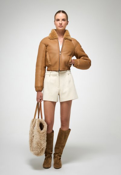Giacca bomber in pelle marrone con collo in pelliccia sintetica, pantaloni corti crema e stivali al ginocchio in suede marrone. Tiene una borsa morbida beige.
