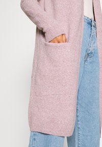 Ljusrosa cardigan med ribbade mudd och sidofickor, tillverkad av ett mjukt material. Matchad med ljusblå jeans med en avslappnad passform.