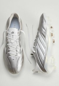 Paire de chaussures de football Adidas argentées avec des lacets blancs, une chaussure vue de dessus et l'autre de côté sur une surface blanche.