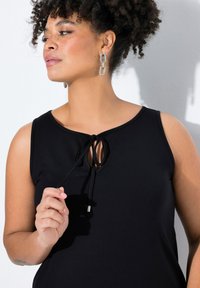 Czarny tank top bez rękawów z okrągłym dekoltem i detalem w postaci zawiązania z przodu. Gładki materiał z subtelnym połyskiem. Modelka nosi srebrne kolczyki w kształcie łańcuszka.