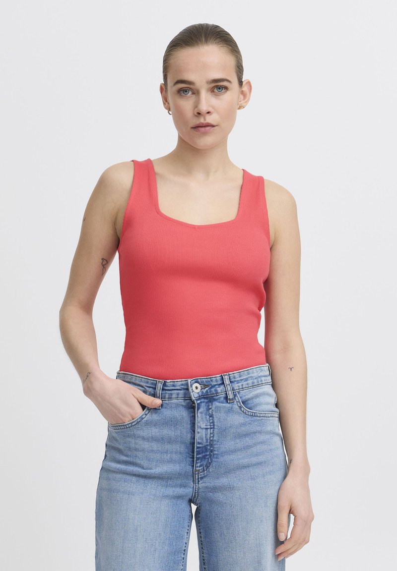Femme à la peau claire et aux cheveux attachés porte un haut sans manches corail et un jean taille haute bleu clair, debout avec une main dans la poche.