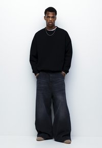 Sort oversize sweatshirt og mørkeblå vidde jeans, med mansjetter hvile på lysfarvede sko. Model står mod en ensfarvet baggrund.