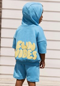 Modrá bavlněná mikina s kapucí a žlutou grafikou "FUN VIBES" na zadní straně, spárovaná s odpovídajícími modrými šortkami. Hladká textura a volný střih.
