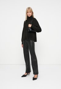 Schwarzer, strukturierter, übergroßer Pullover mit hohem Kragen, kombiniert mit schwarzen Ledergeraden Hosen und spitz zulaufenden Absätzen. Kleine schwarze Handtasche als Akzent.