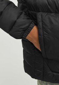 Crna puffer jakna prikazuje quilted teksturu s elastičnim manžetama. Ruka se pruža prema bočnoj džepu, ističući funkcionalnost odjevnog predmeta.