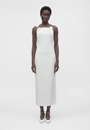 Žena v bielych midi šatách bez rukávov s kvetinovými zdobeniami na ramienkach, nosí biele lodičky s remienkami okolo členkov.