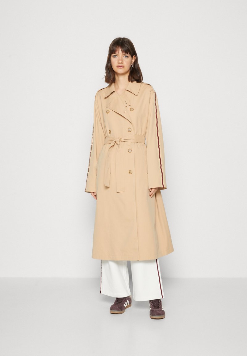 Trench coat beige con botones de doble fila, cintura ceñida con cinturón y rayas laterales rojas; combinado con pantalones anchos blancos y zapatillas marrones.