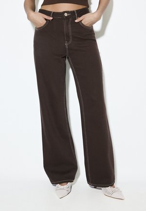 Brune jeans med høj talje og brede ben med hvide syninger, båret med hvide spidse flettede hæle, hænderne i forlommerne.