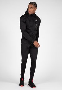 Zwarte rits-hoodie met een slim fit ontwerp, textuurstof en verstelbare trekkoorden. Gecombineerd met zwarte slim-fit joggers en sportschoenen.