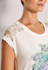 Vrouw die een wit shirt draagt met kanten schouderpanelen en een kleurrijk abstract patroon aan de voorkant, afgebeeld van de nek tot halverwege de arm.