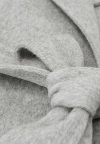 Siva filcna tkanina z mehko teksturo, z značilnostjo vozlanega okrasnega traku. Dizajn vključuje večplastne naborke, ki poudarjajo globino.