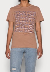 Una persona está de pie vistiendo una camiseta de color marrón claro con un diseño gráfico geométrico, combinada con unos vaqueros rotos de color azul claro.