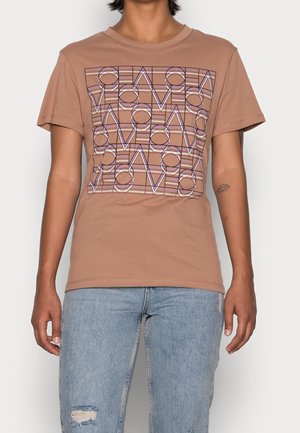 Een persoon staat in een lichtbruine T-shirt met een geometrisch grafisch ontwerp, gecombineerd met lichtblauwe ripped jeans.