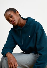Dark teal hooded sweatshirt with a drawstring, featuring a small logo on the chest. The model sits in gray trousers, showcasing a relaxed fit.