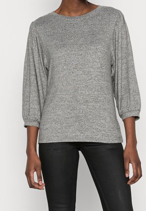 Kobieta ubrana w szarą bluzkę na długi rękaw z marszczonymi mankietami i czarne dopasowane spodnie, stojąca na jednolitym tle.