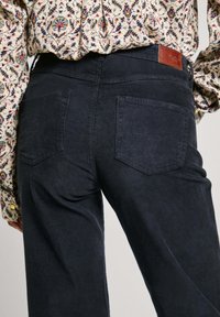 Pantalones de pana azul oscuro con una superficie texturizada, que cuentan con dos bolsillos traseros y un parche de cuero marrón de la marca en la cinturilla.