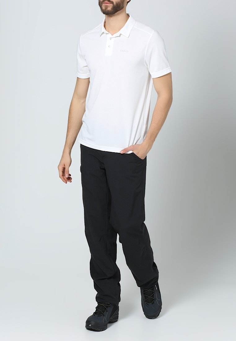 Homme portant un polo blanc à manches courtes, un pantalon noir et des chaussures de randonnée noires, debout avec une main dans la poche.