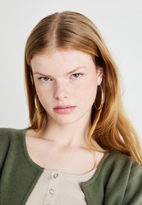 Πράσινο πλεκτό μπολερό με στρογγυλή λαιμόκοψη, τοποθετημένο πάνω από ένα ανοιχτό μπεζ ριμπ μπλουζάκι με κουμπιά σνάπ. Μεγάλες χρυσές κρίκοι σκουλαρίκια.