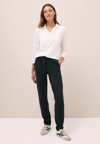 Bílá polokošile s dlouhým rukávem a límečkem, spárovaná s tmavě zelenými joggingovými kalhotami s elastickým pasem a nastavitelnými šňůrkami.