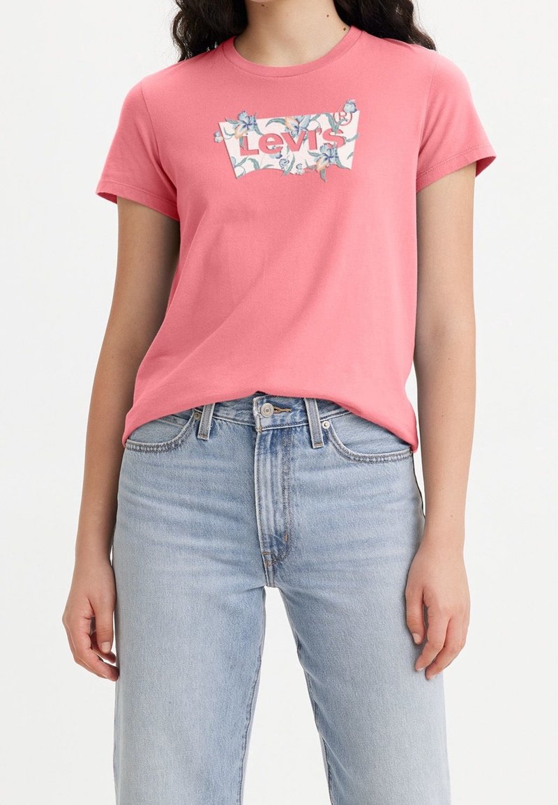 Femme portant un t-shirt Levi's rose avec un logo floral et un jean taille haute bleu clair, debout les mains détendues le long du corps.