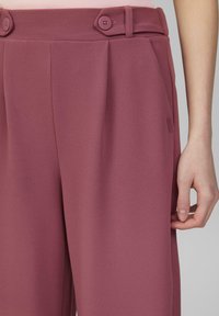 Pantalon plissé taille haute en tissu mauve, doté de deux accents de boutons et de poches latérales. Texture lisse et coupe sur mesure.