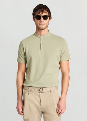 Polo a maniche corte verde chiaro con collo alto e patta con tre bottoni, abbinata a pantaloni beige con cintura e fibbia argentata.